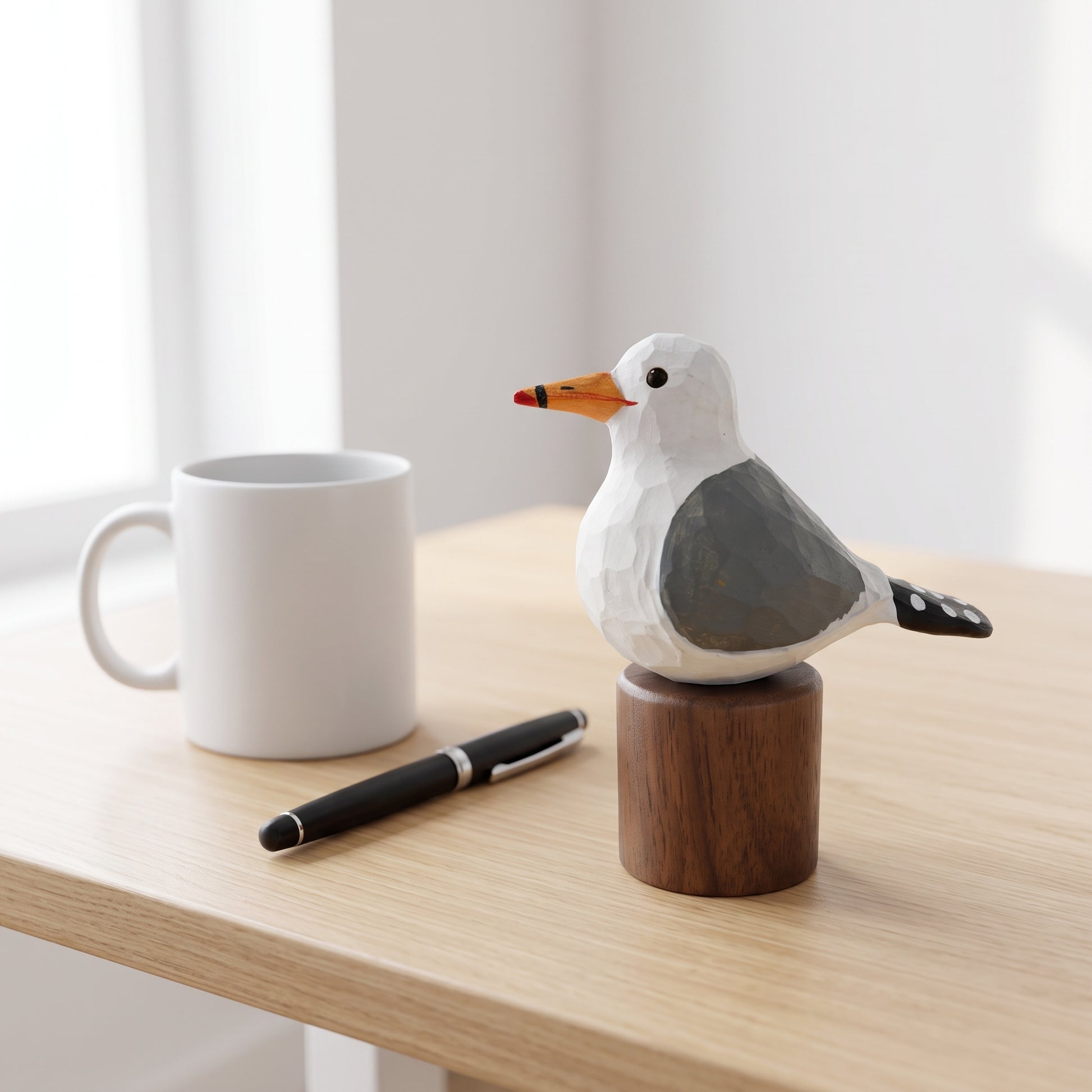 A hand-carved wooden California gull figurine perched on a cylindrical wooden base, displayed on a light wood desk next to a white ceramic mug and a black pen.