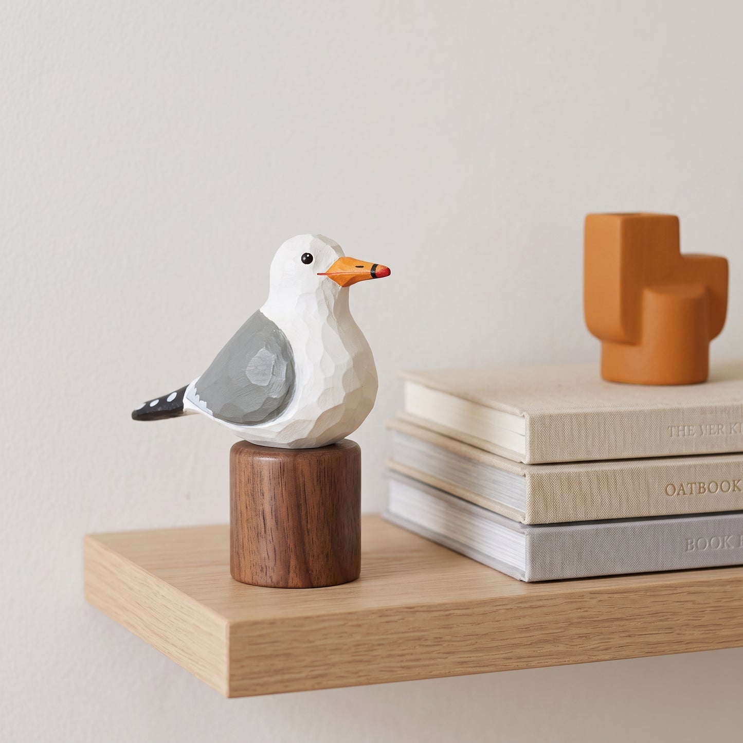 A geometric California gull figurine with a white head, gray wings, and an orange beak, mounted on a round wooden base and displayed on a light wood shelf next to stacked books.