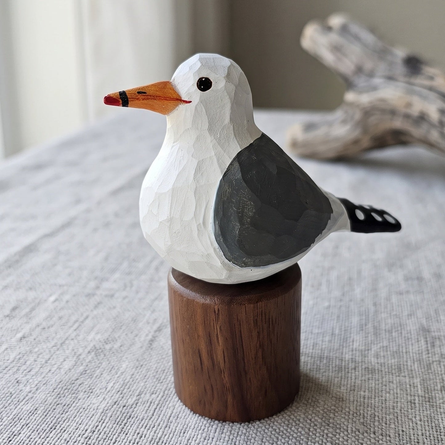A hand-carved wooden California gull figurine with a white head, gray wing, orange beak, and black eye, mounted on a small round wooden base. The figurine is placed on a textured light gray fabric surface with driftwood and a window in the background.