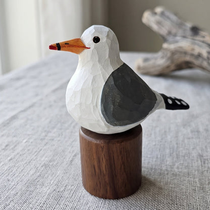 A hand-carved wooden California gull figurine with a white head, gray wing, orange beak, and black eye, mounted on a small round wooden base. The figurine is placed on a textured light gray fabric surface with driftwood and a window in the background.