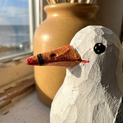 A close-up of a handcrafted wooden California gull figurine with a white-painted body and a reddish-brown beak, positioned in front of a large tan ceramic vase on a windowsill.