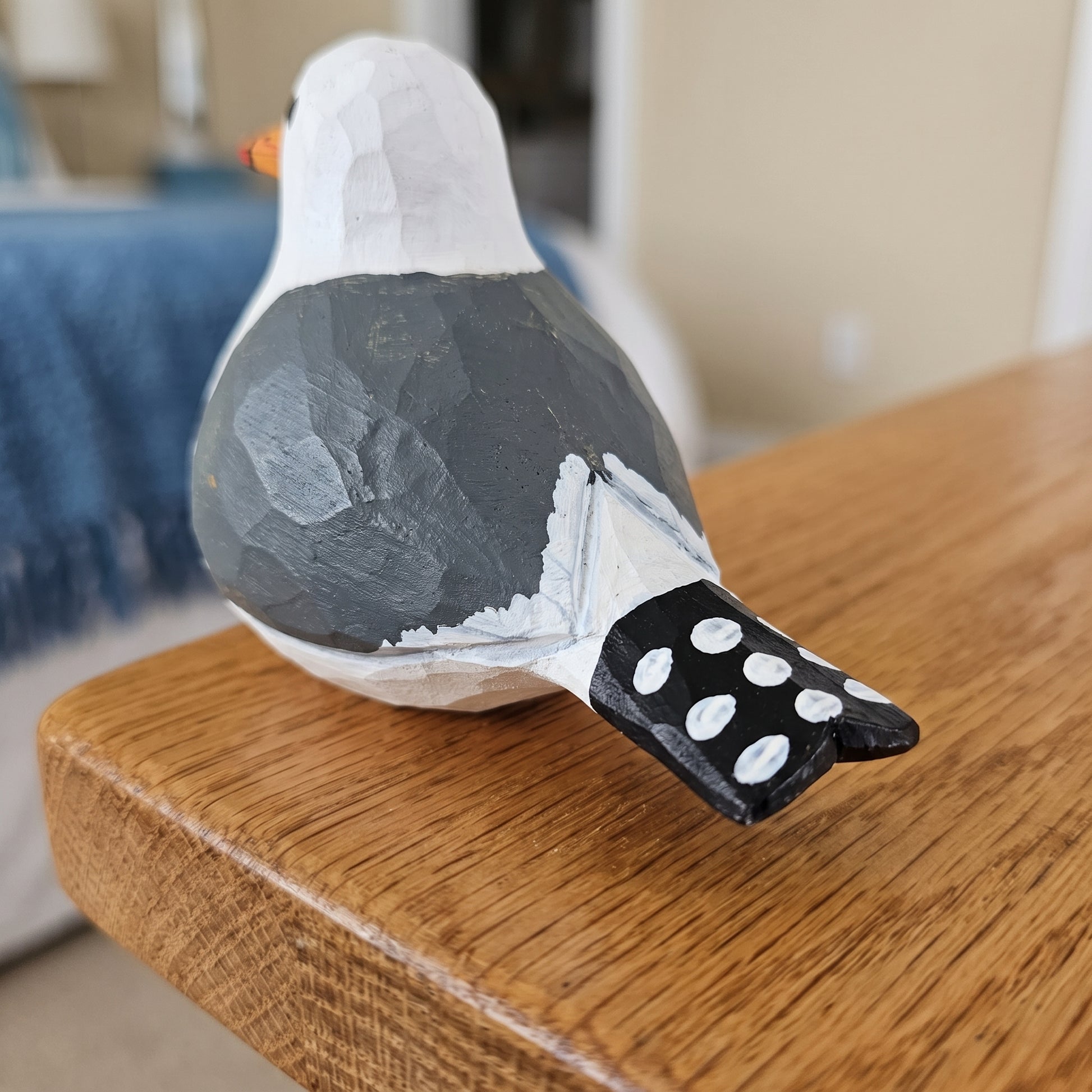 A hand-painted wooden California gull figurine with a white head, dark gray back, and black tail with white spots, resting on a wooden table in a home setting.