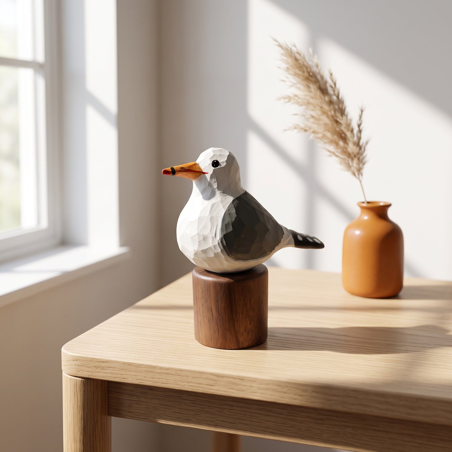 A geometric California gull figurine with a white, gray, and orange body mounted on a round wooden base, displayed on a light wood table in a sunlit room next to a small terracotta vase with dried grass.