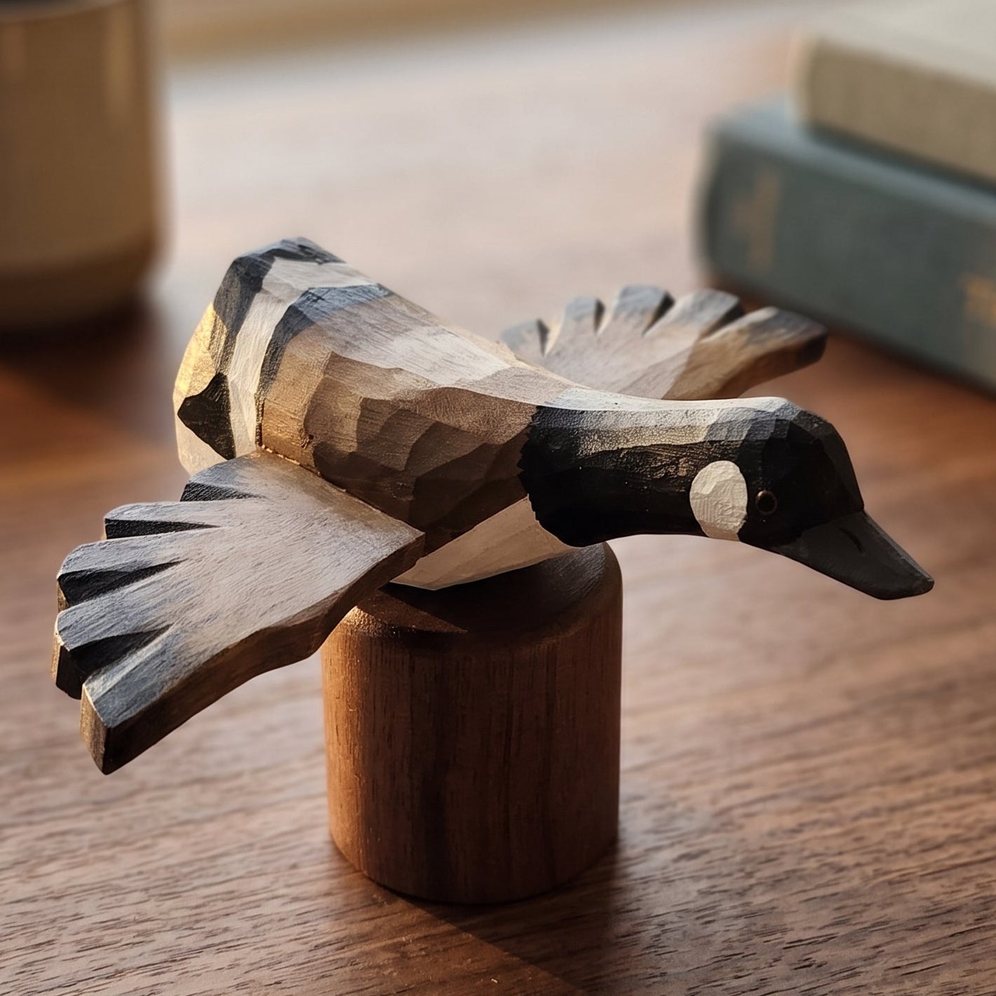 A hand-carved wooden Canada Goose Flying figurine perched on a small wooden block, displayed on a wooden table with a blurred coffee mug and books in the background.