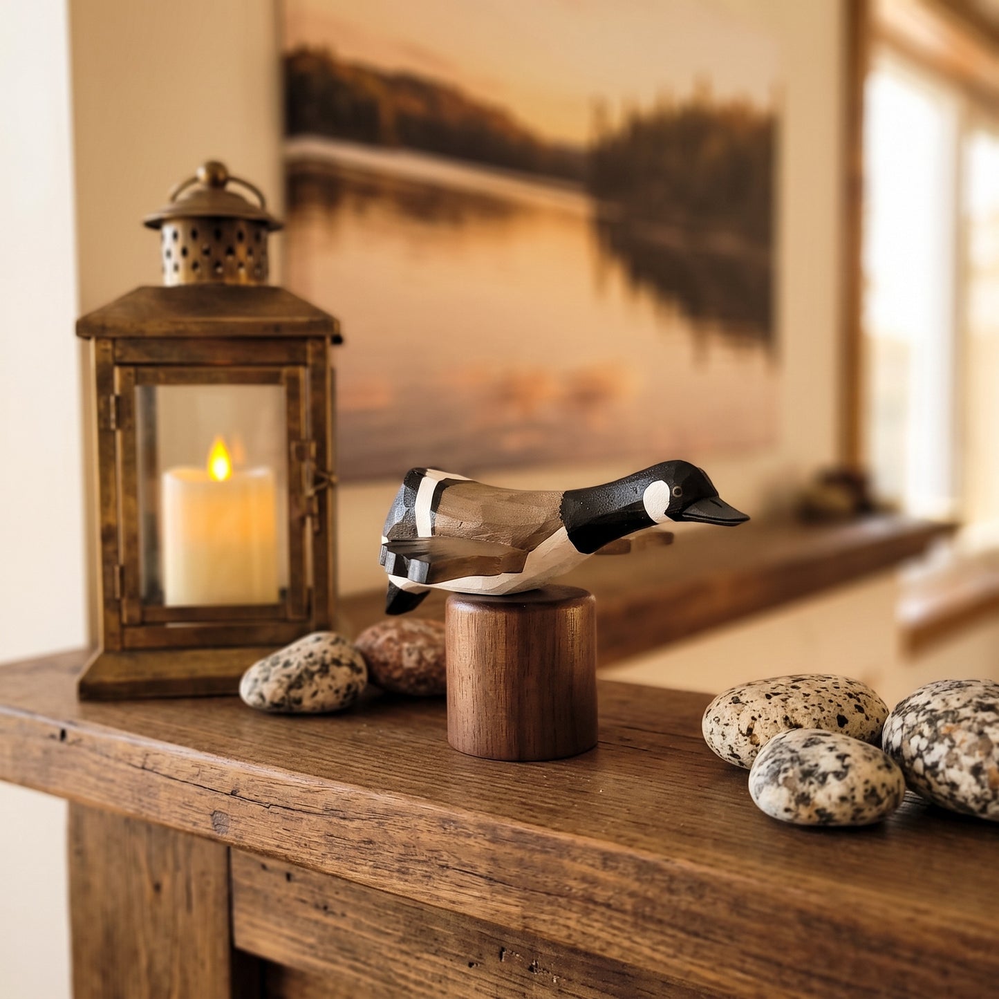 A carved wooden Canada Goose Flying deco displayed on a small wooden stand on a rustic wooden table, with a lit lantern, decorative stones, and a warm-toned framed landscape painting in the background.