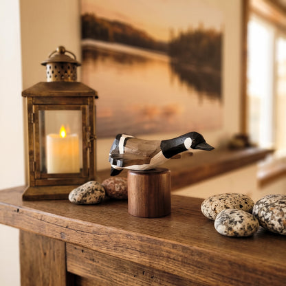 A carved wooden Canada Goose Flying deco displayed on a small wooden stand on a rustic wooden table, with a lit lantern, decorative stones, and a warm-toned framed landscape painting in the background.