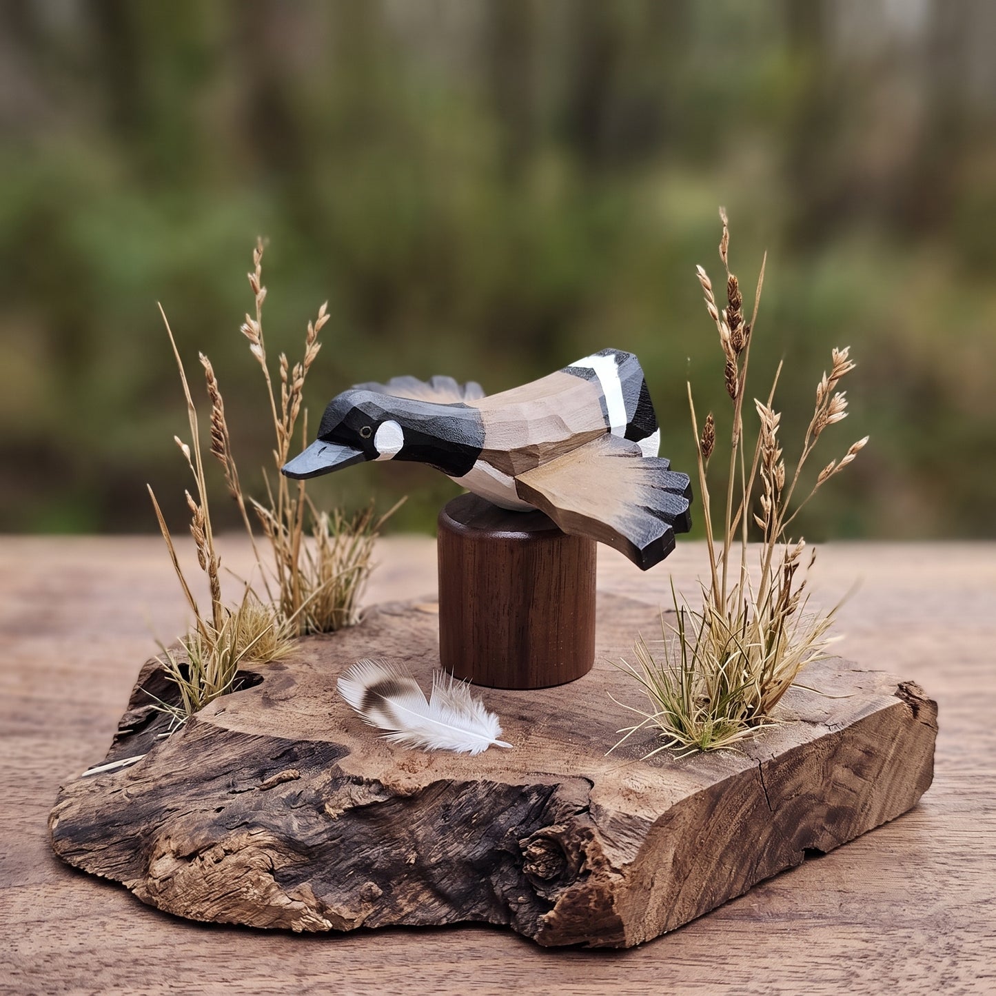 A handcrafted wooden Canada Goose Flying figurine with geometric facets, mounted on a dark wooden cylinder and displayed on a rustic live-edge wood slab with dried grasses and a small feather, set against a blurred outdoor background.