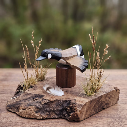 A handcrafted wooden Canada Goose Flying figurine with geometric facets, mounted on a dark wooden cylinder and displayed on a rustic live-edge wood slab with dried grasses and a small feather, set against a blurred outdoor background.