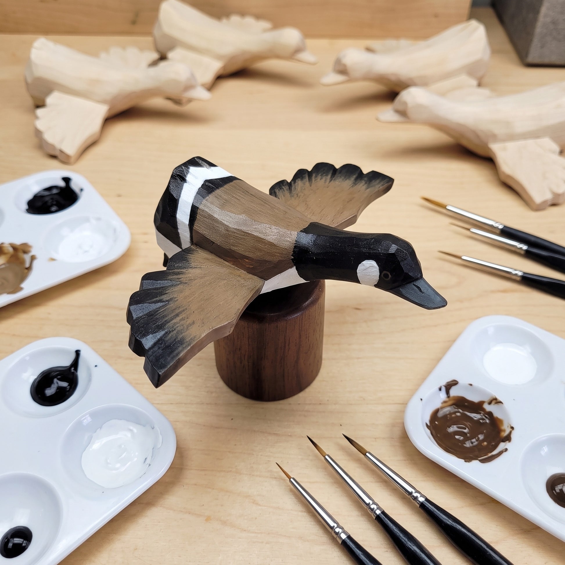A handcrafted wooden Canada Goose Flying figurine with black, brown, and white painted details sits on a wooden stand on a light wood table, surrounded by paint palettes, paintbrushes, and unfinished wooden duck shapes.