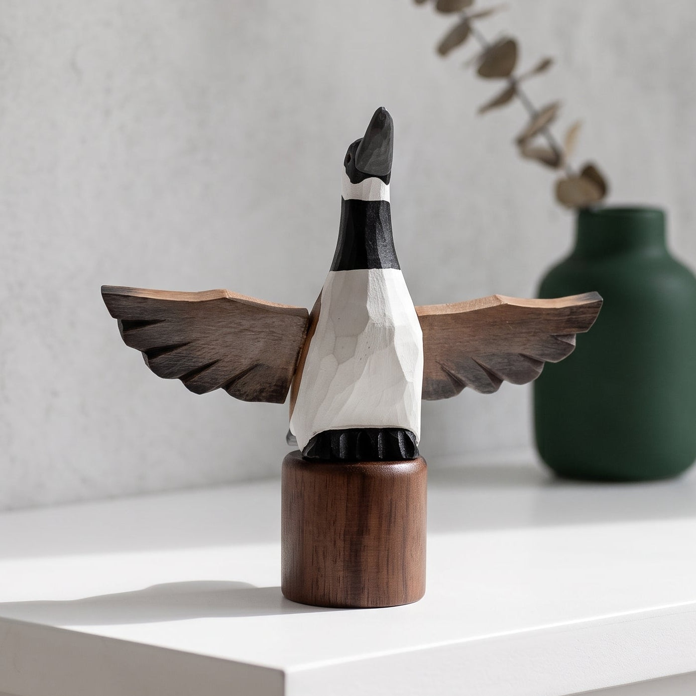 A handcrafted wooden Canada Goose Flying figurine with outstretched wings, mounted on a cylindrical wooden base, displayed on a white shelf next to a green ceramic vase.