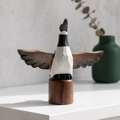 A handcrafted wooden Canada Goose Flying figurine with outstretched wings, mounted on a cylindrical wooden base, displayed on a white shelf next to a green ceramic vase.