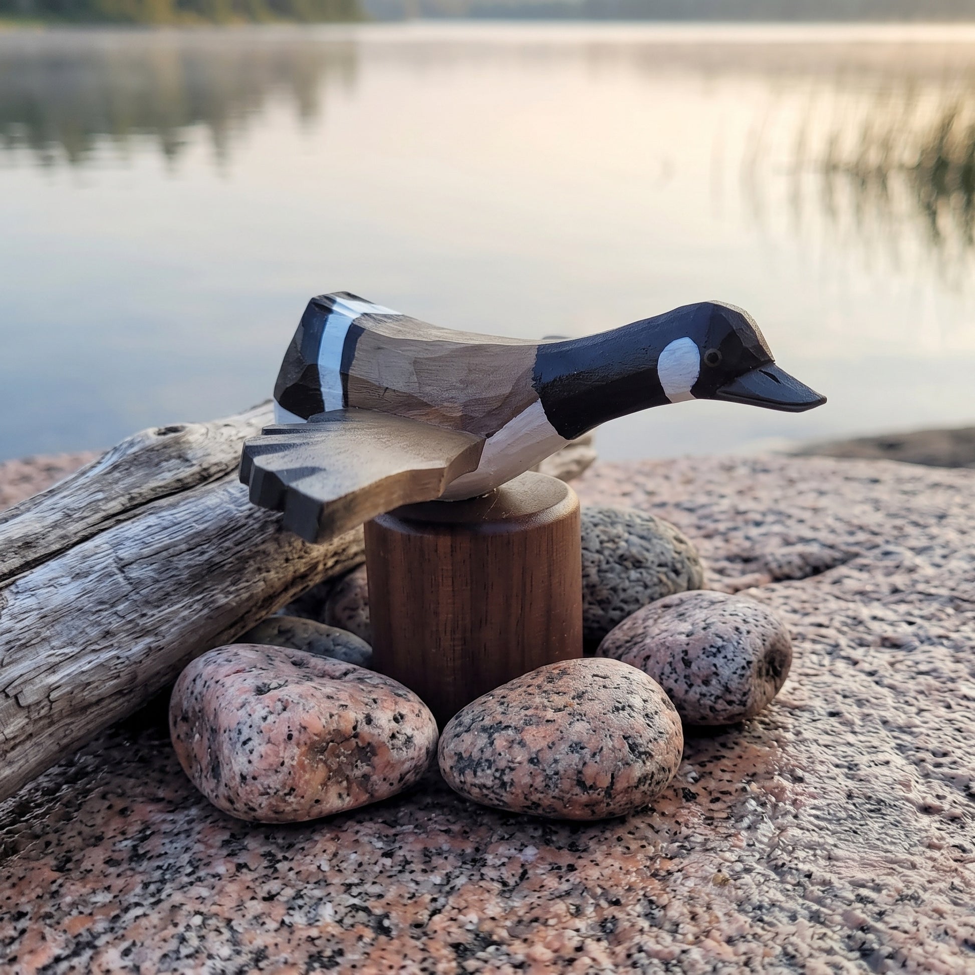 A carved wooden Canada Goose Flying figurine with a black, white, and tan body, displayed on a small wooden pedestal outdoors on a rock beside a log and lake in the background.