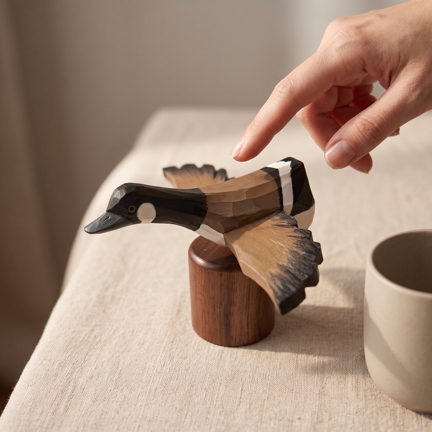 A hand points toward a carved wooden Canada Goose Flying figurine perched on a small wooden base, set on a beige fabric-covered table with a neutral cup nearby.