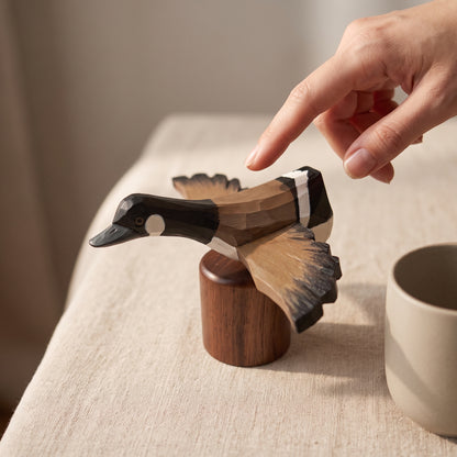 A hand points toward a carved wooden Canada Goose Flying figurine perched on a small wooden base, set on a beige fabric-covered table with a neutral cup nearby.