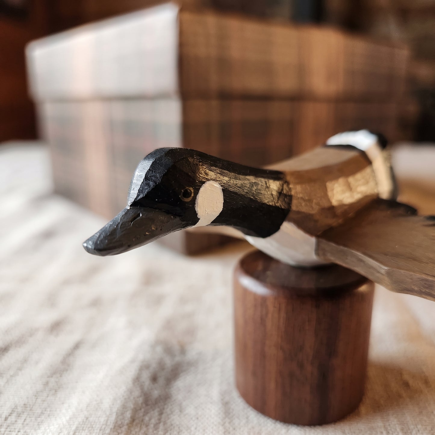 A hand-carved wooden Canada Goose Flying figurine with a black, white, and brown painted head, mounted on a cylindrical wooden base. The background is softly blurred, showing a plaid box and a neutral fabric surface.