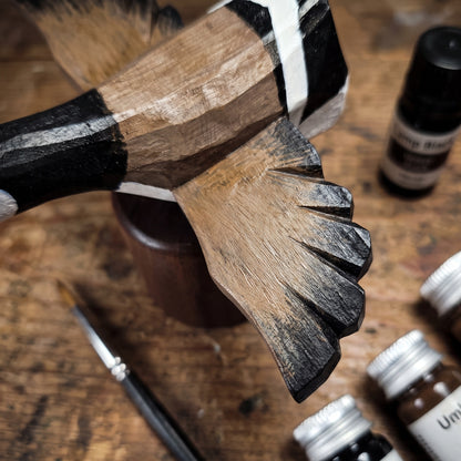 A close-up of a hand-carved wooden Canada Goose Flying figurine with black, white, and natural wood tones, resting on a rustic wooden table with paint bottles and a paintbrush nearby.