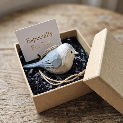 A small Canada jay bird-shaped ornament in pale blue and cream sits in a kraft gift box with black shredded paper filler and a card that reads 'Especially For You.' The box is open on a wooden surface.