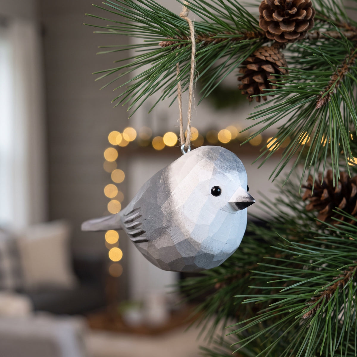 A geometric Canada jay bird-shaped Christmas ornament hanging from a pine tree branch, with blurred warm lights and a cozy indoor background.