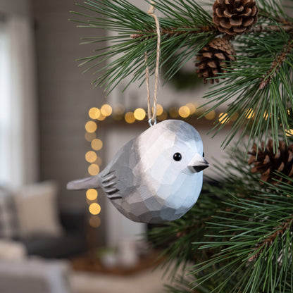 A geometric Canada jay bird-shaped Christmas ornament hanging from a pine tree branch, with blurred warm lights and a cozy indoor background.