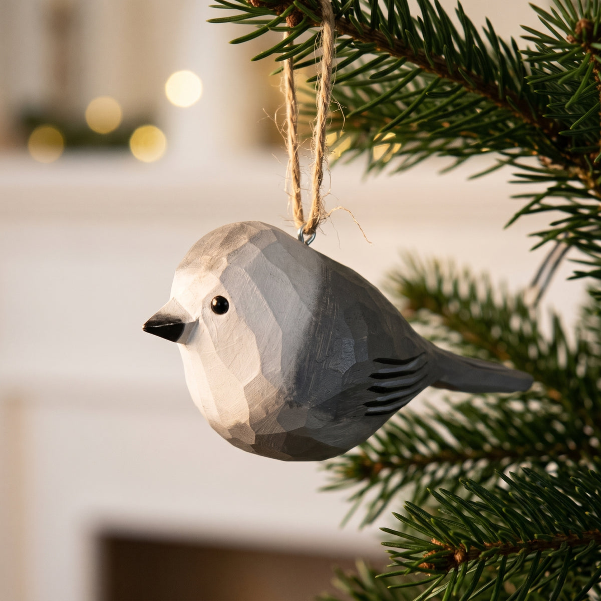 A geometric, faceted wooden Canada jay Christmas ornament in light gray and white, hanging from a natural jute string on a green pine tree branch with a blurred indoor background.