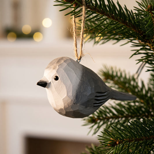 A geometric, faceted wooden Canada jay Christmas ornament in light gray and white, hanging from a natural jute string on a green pine tree branch with a blurred indoor background.