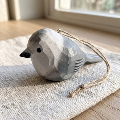 A carved wooden Canada jay bird ornament with a natural wood twine loop, resting on a textured linen cloth on a wooden table near a window.