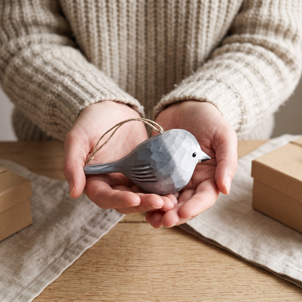 A person in a beige knit sweater holds a small faceted Canada jay bird ornament in their hands over a wooden table with linen cloths and gift boxes.