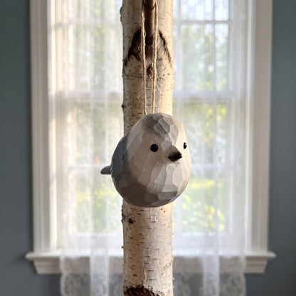 A low-poly Canada jay bird hanging ornament made from birch wood, suspended by a jute string in front of a window with sheer white curtains.