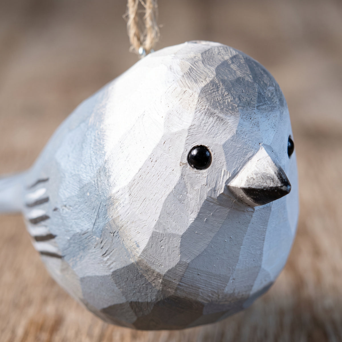 A close-up of a carved wooden Canada jay bird ornament with a geometric, faceted design, painted in shades of gray and white, hanging by a natural jute twine string.