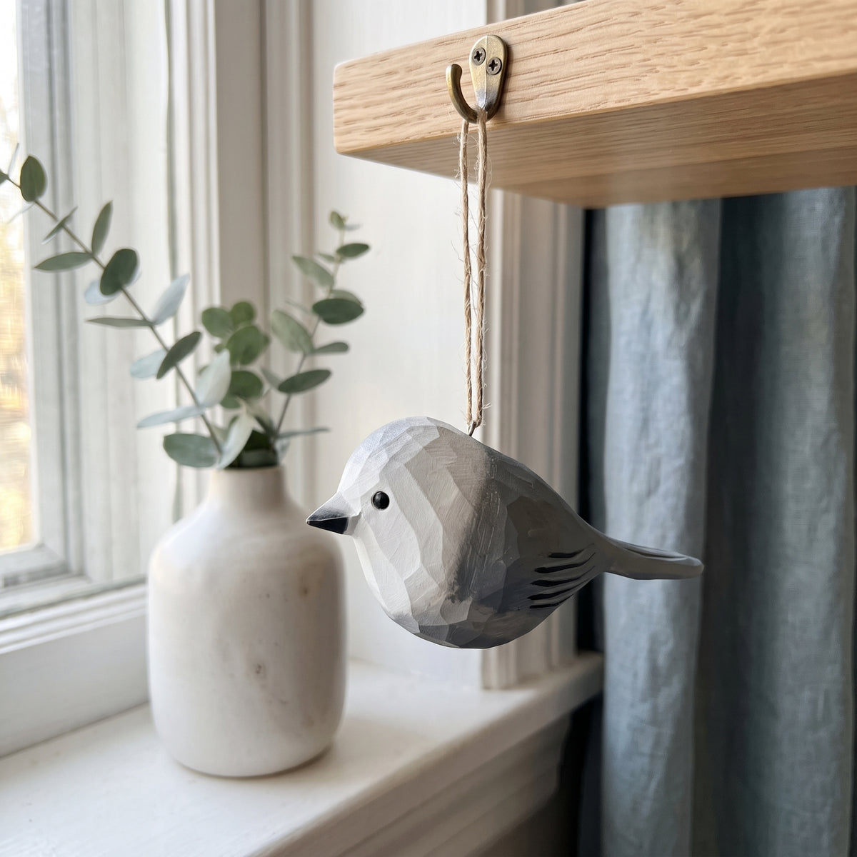 A geometric Canada jay  bird-shaped wooden ornament with a faceted polygonal design, hanging from a wooden shelf hook with a twine string. The ornament is styled on a windowsill next to a white ceramic vase with eucalyptus branches and a blue-gray curtain.