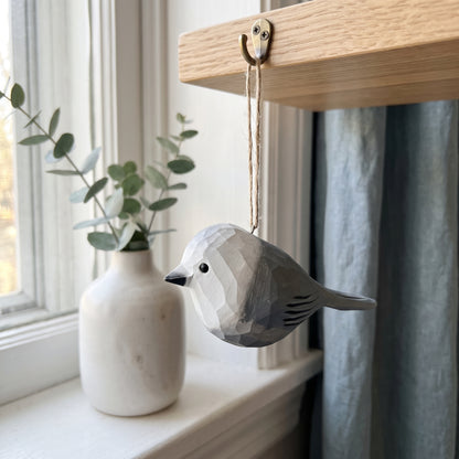 A geometric Canada jay  bird-shaped wooden ornament with a faceted polygonal design, hanging from a wooden shelf hook with a twine string. The ornament is styled on a windowsill next to a white ceramic vase with eucalyptus branches and a blue-gray curtain.