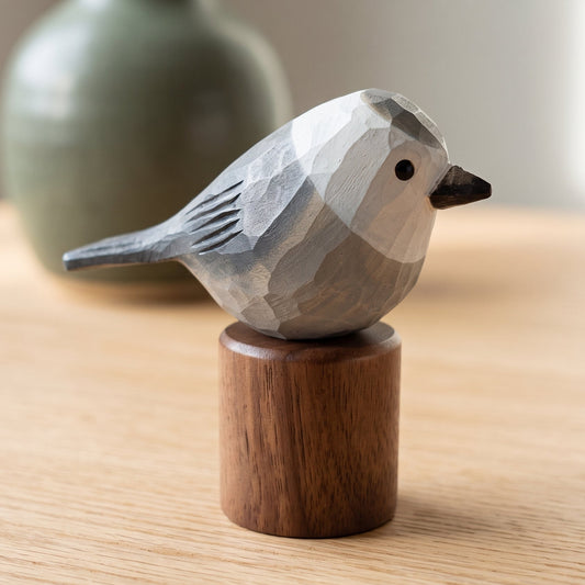 A small carved Canada jay figurine with a gray and white geometric body, black beak, and dark gray wing, mounted on a round brown wooden base and photographed on a light wood tabletop with a blurred green vase in the background.