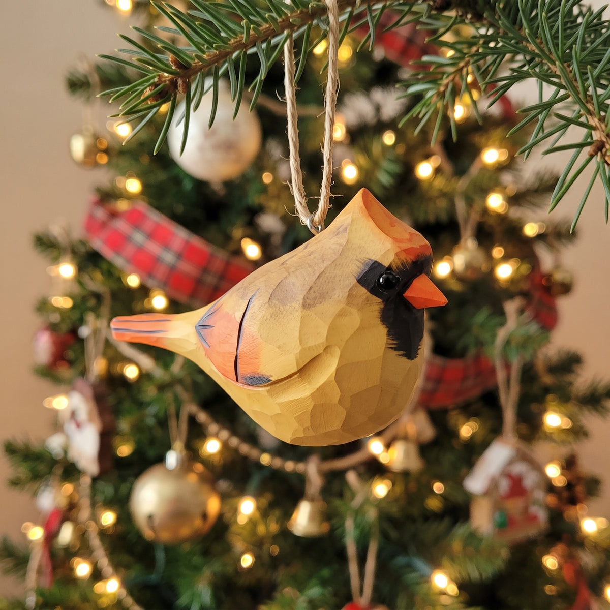 A carved wooden cardinal Christmas ornament hanging from a pine branch in front of a lit Christmas tree.
