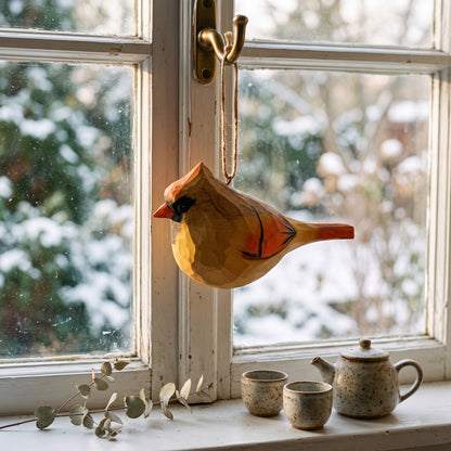 A hand-painted wooden cardinal ornament hangs from a brass hook on a window frame, with a snowy outdoor view in the background and a ceramic teapot and cups on the windowsill.