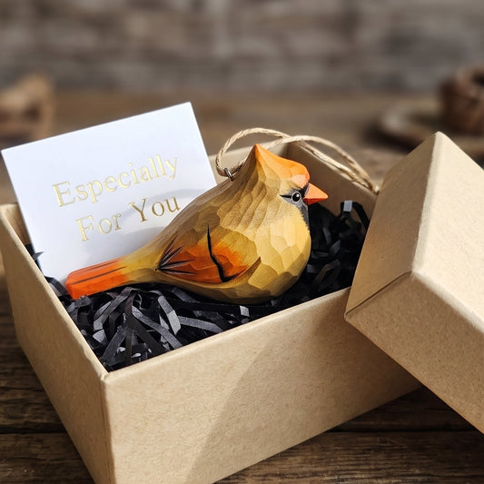 A carved wooden cardinal ornament with a gold and orange finish rests in black shredded paper inside an open kraft gift box, with a white card reading 'Especially For You' and the box lid leaning beside it.