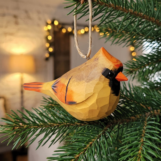 A handcrafted wooden cardinal bird ornament with a natural jute hanging loop, displayed on a green pine Christmas tree branch with warm bokeh lights in the background.