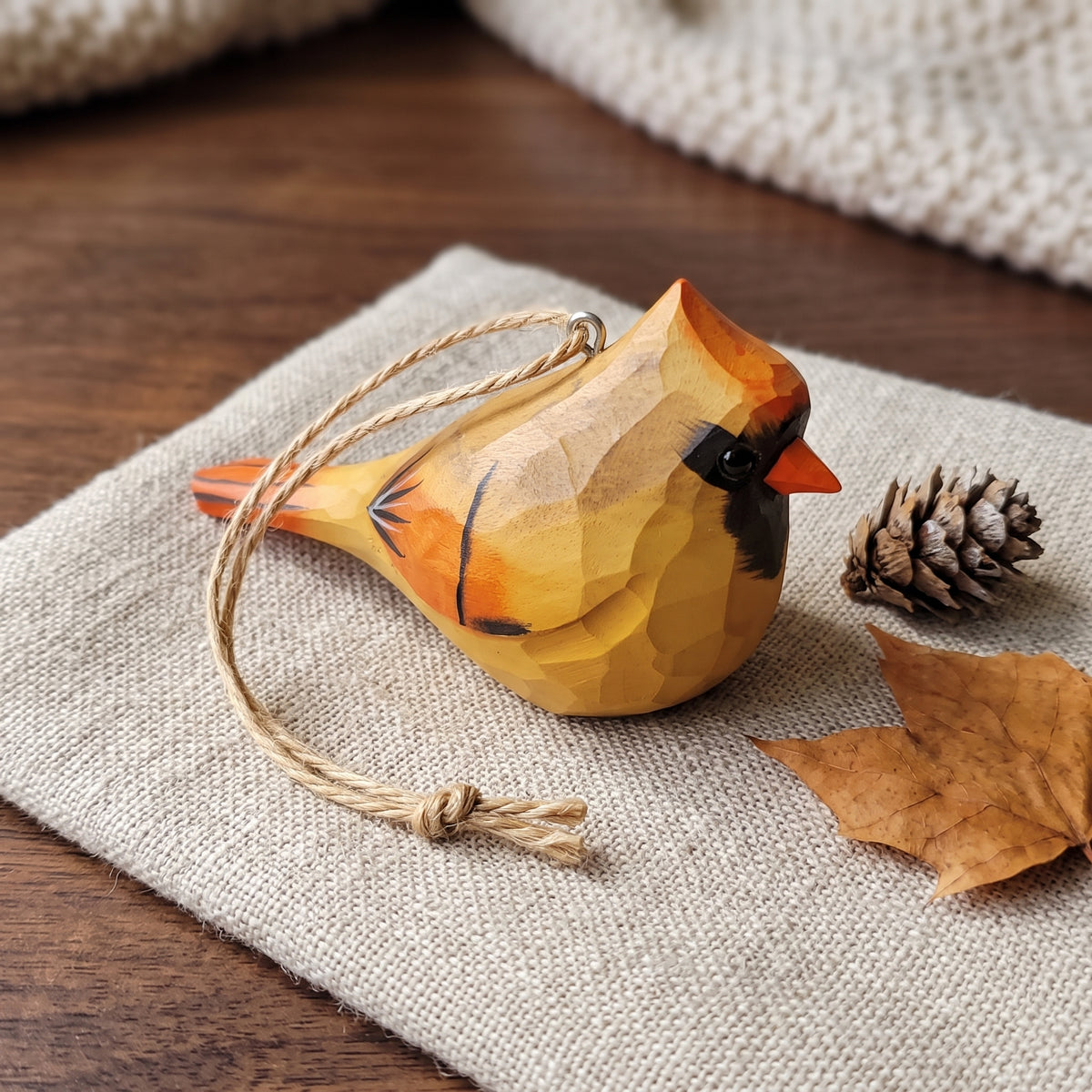 A carved wooden cardinal ornament with a geometric faceted design, painted in warm yellow, orange, and black, hanging from a natural jute rope. It is styled on a textured beige linen cloth next to a pinecone and a dried maple leaf on a dark wood surface.