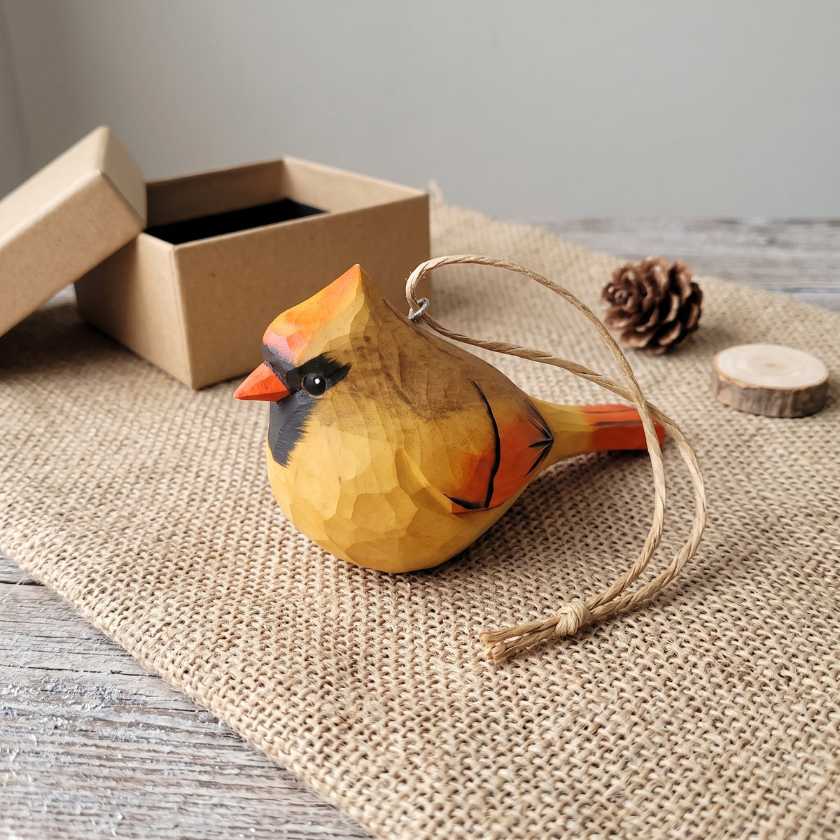 A carved wooden cardinal bird ornament with a natural wood grain finish, featuring a hanging twine loop, displayed on a burlap mat with a pinecone, wood slice, and gift box in the background.