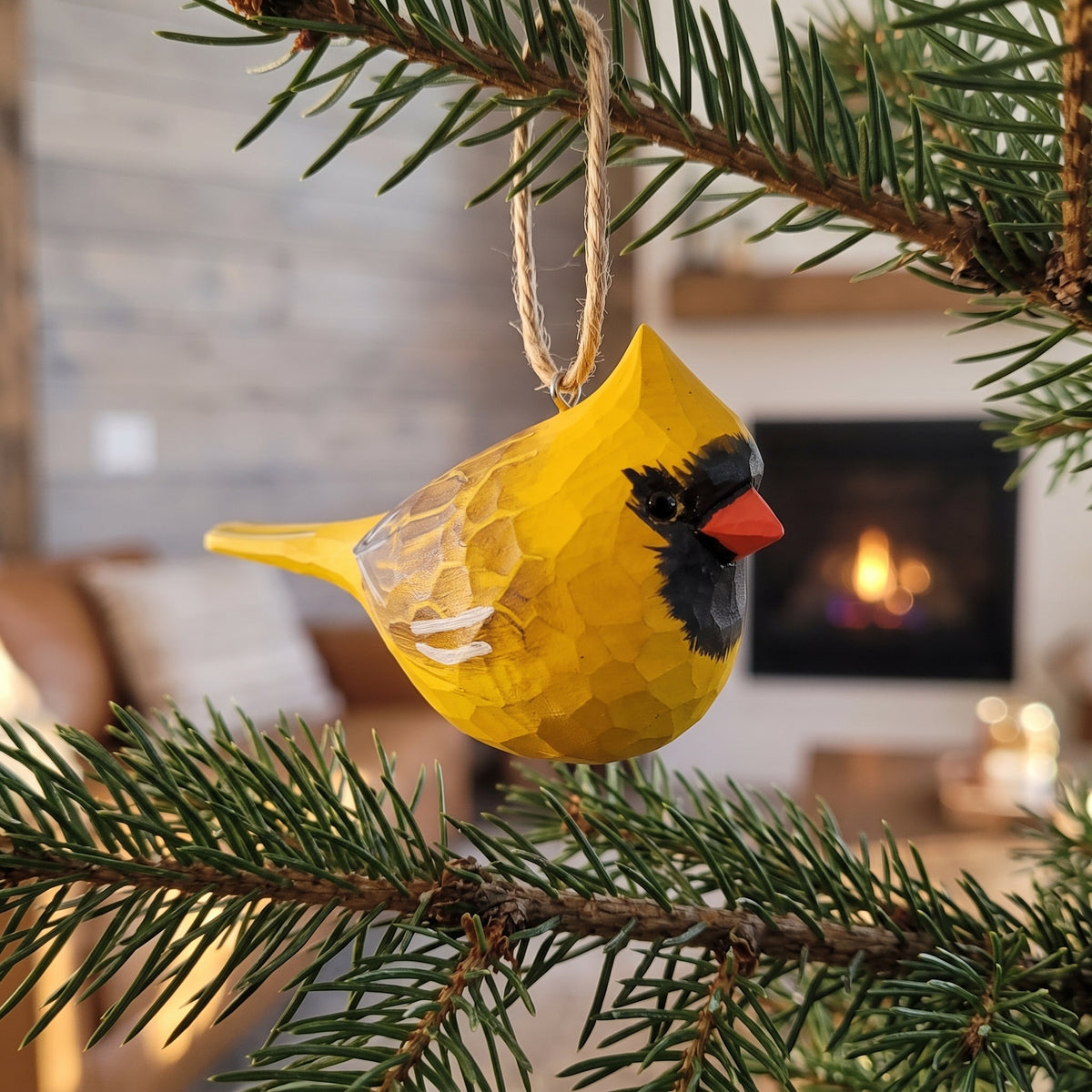 A yellow cardinal bird Christmas ornament with a textured honeycomb finish, hanging from a natural twine string on a pine tree branch in front of a cozy living room background with a fireplace.