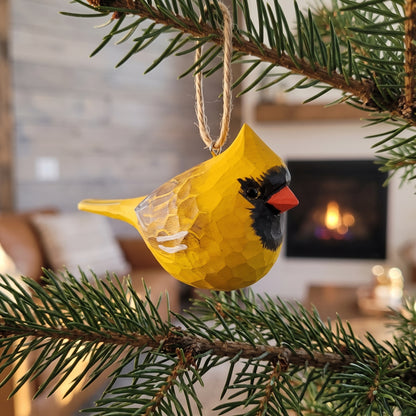 A yellow cardinal bird Christmas ornament with a textured honeycomb finish, hanging from a natural twine string on a pine tree branch in front of a cozy living room background with a fireplace.
