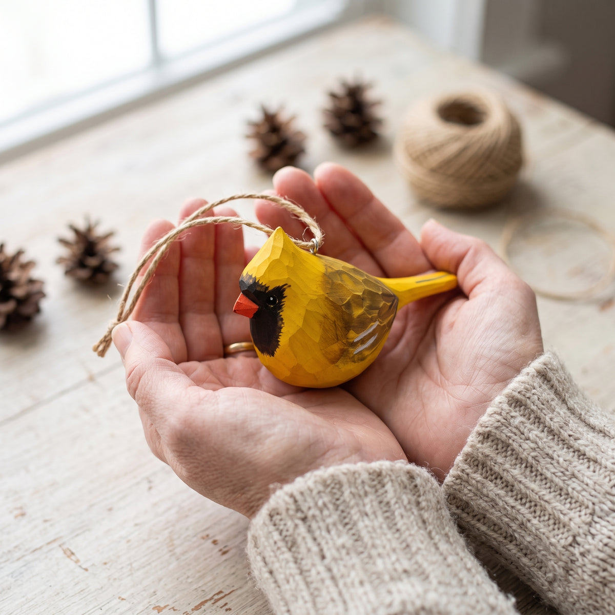 A person holds a small yellow bird ornament in their hands. The ornament has a black face, red beak, and a hanging twine loop.