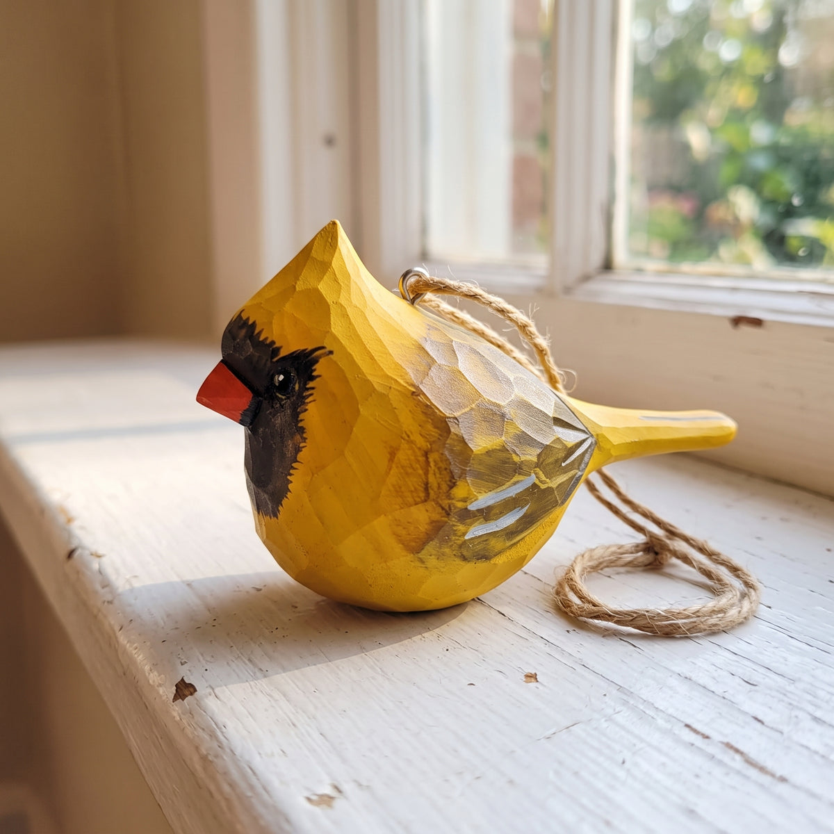 A small wooden cardinal bird ornament with a faceted carved texture, painted in bright yellow with a black face, red beak, and natural jute hanging twine, resting on a white-painted wooden windowsill.
