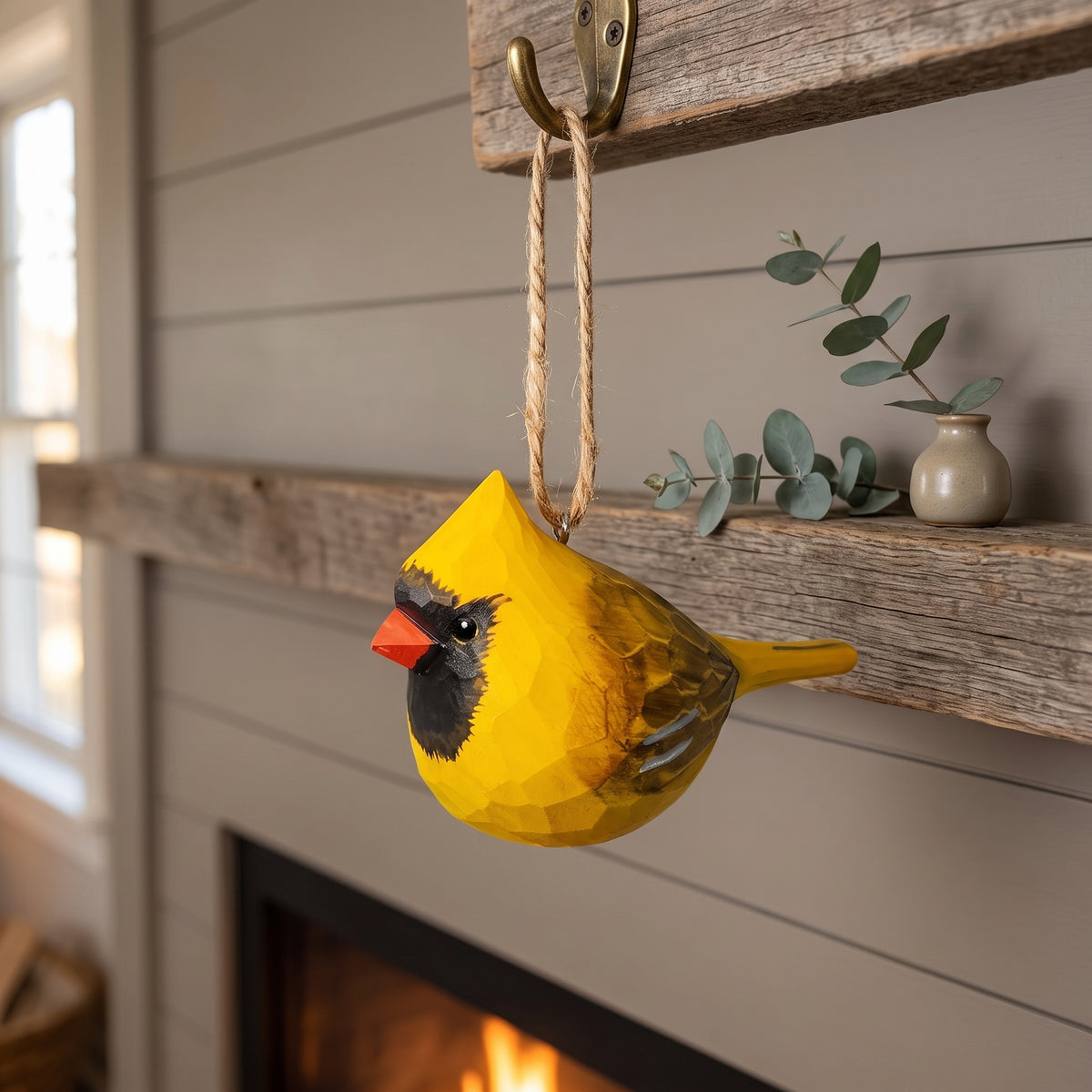 A yellow cardinal-shaped wall hanging with a rope loop hangs from a rustic wooden hook on a fireplace mantel. The bird ornament has a bright yellow body, a black face, and a red beak, with a small gray branch and ceramic vase on the mantel behind it.