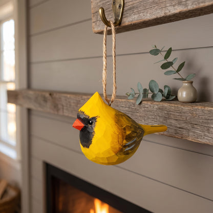 A yellow cardinal-shaped wall hanging with a rope loop hangs from a rustic wooden hook on a fireplace mantel. The bird ornament has a bright yellow body, a black face, and a red beak, with a small gray branch and ceramic vase on the mantel behind it.