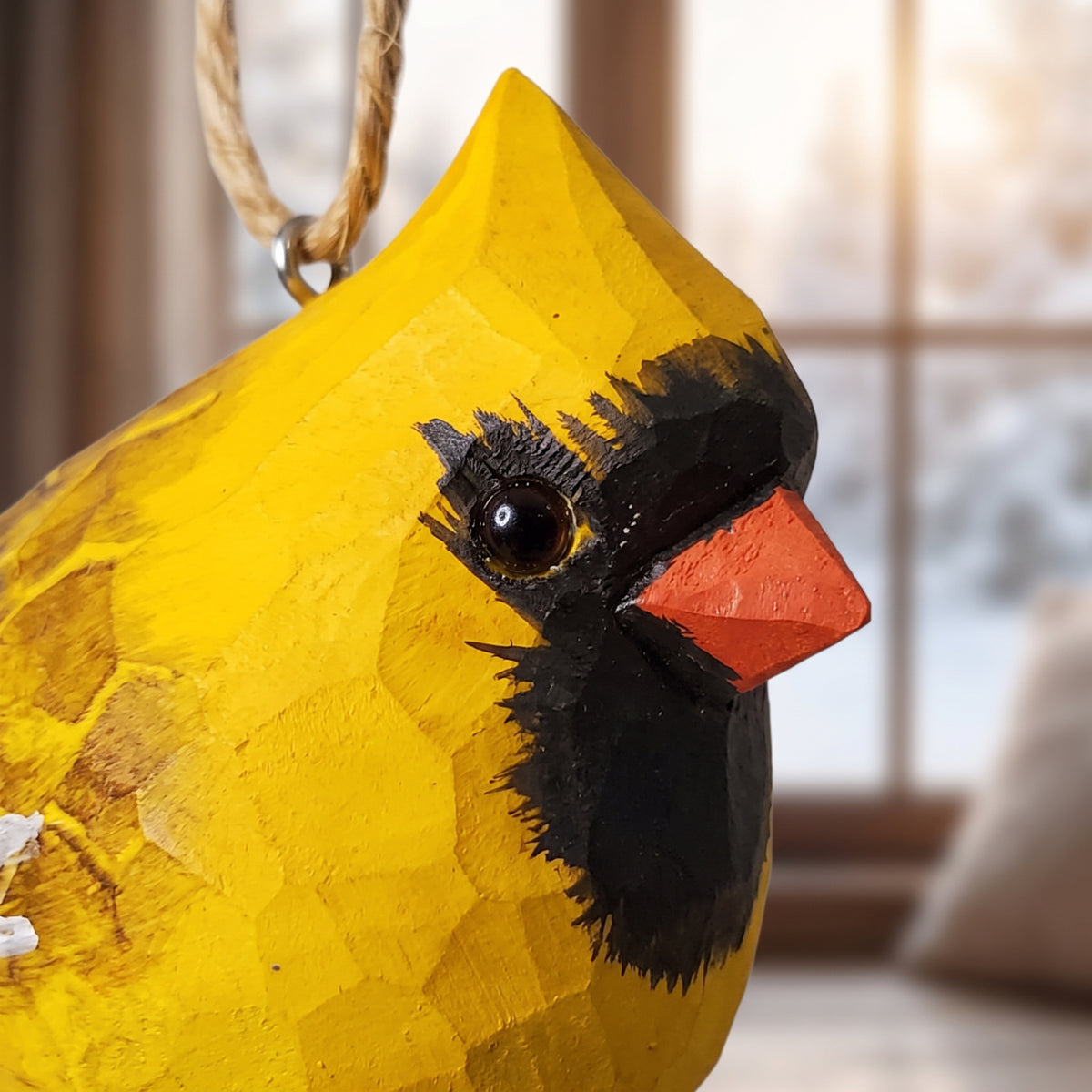 A close-up of a carved wooden cardinal ornament, featuring a bright yellow body, black face, and orange beak, with a hanging loop at the top. The background shows a softly blurred indoor window scene.