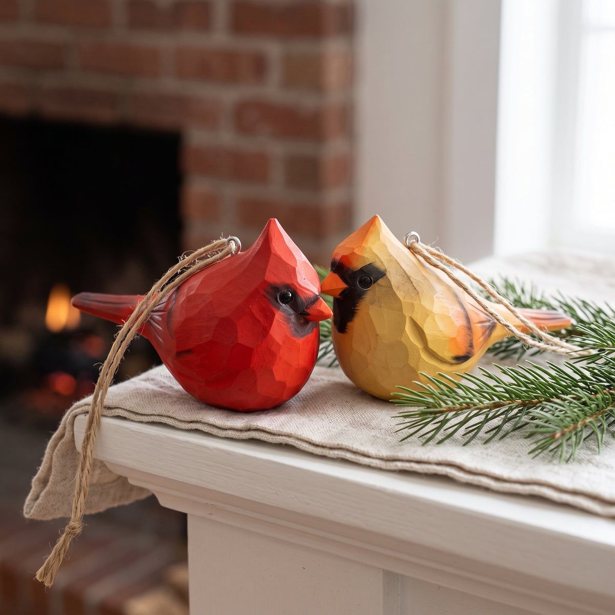 Two carved wooden cardinal bird ornaments, one red and one yellow, resting on a white mantel with a beige cloth, evergreen sprigs, and a blurred fireplace in the background.
