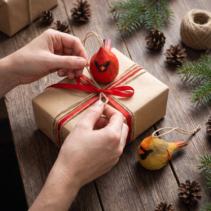 Hands hanging a small red cardinal Christmas ornament on a kraft paper gift box tied with a red ribbon, with pinecones, evergreen branches, twine, and another yellow cardinal ornament on a rustic wooden table.