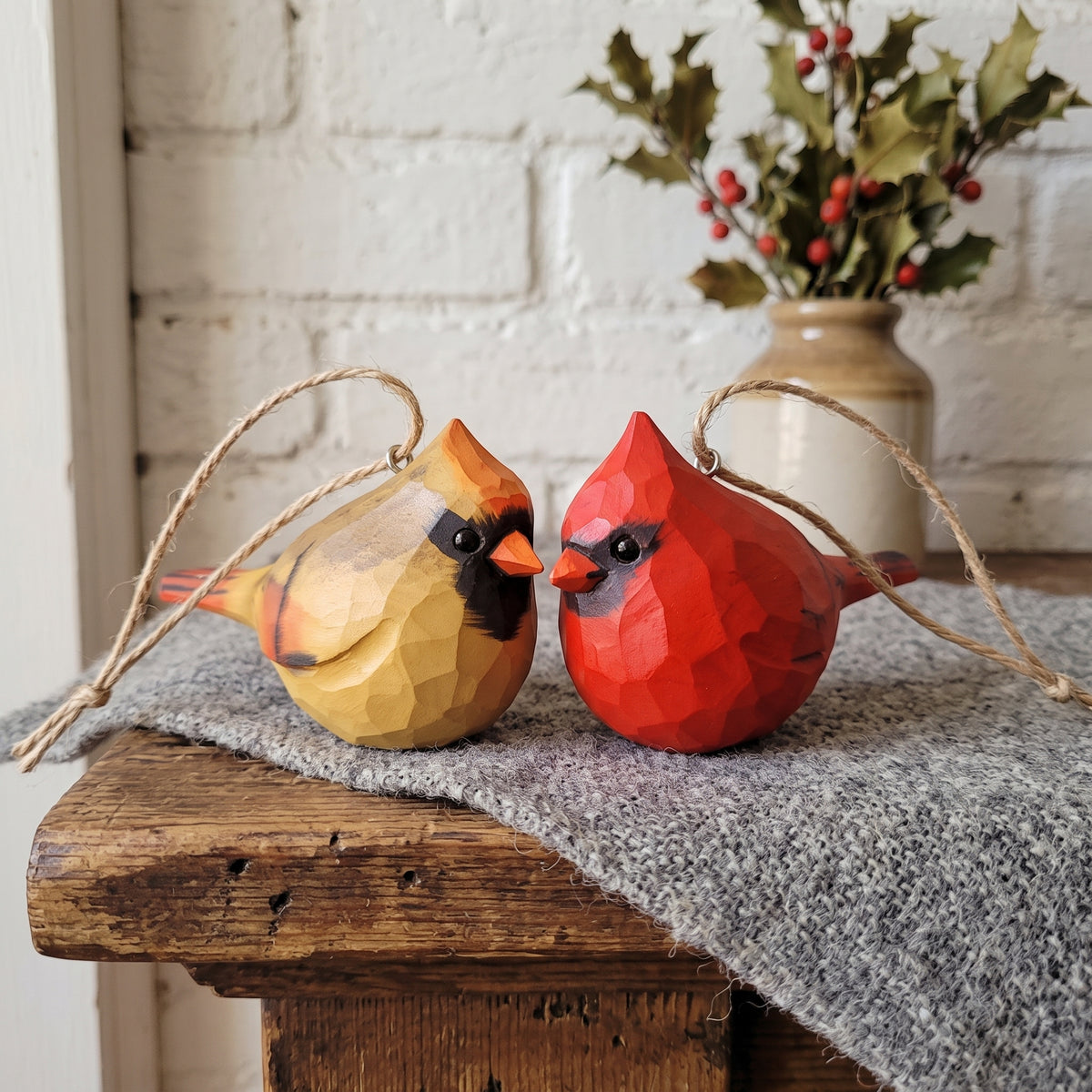 Two geometric wooden cardinal ornaments, one yellow and one red, leaning against each other on a gray knit blanket draped over a rustic wooden table, with a white brick wall and a vase of holly in the background.