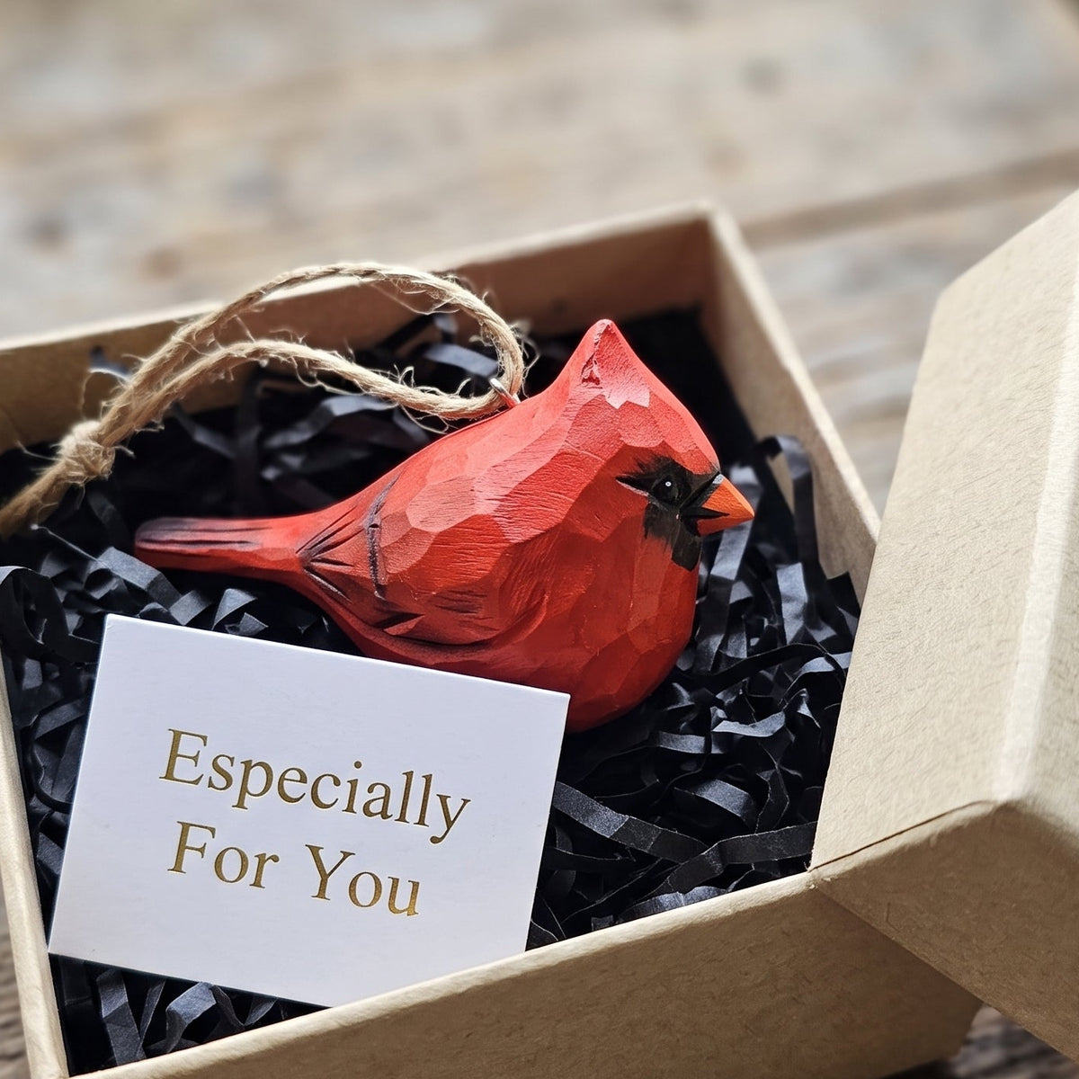 A red cardinal-shaped ornament nestled in black shredded paper inside a kraft gift box, with a small card reading 'Especially For You' on a rustic wooden surface.