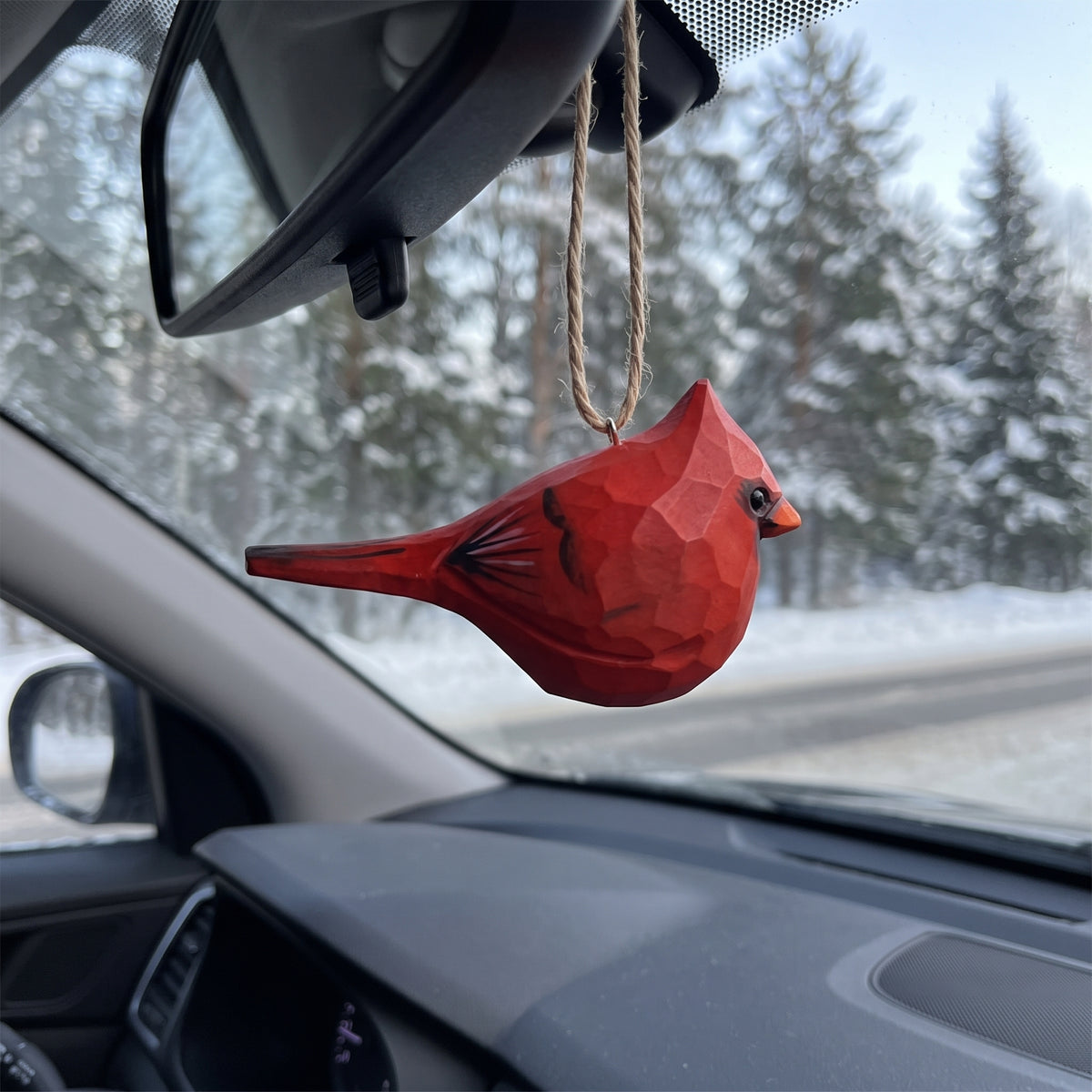 A red geometric bird-shaped car air freshener hanging from a car's rearview mirror, with a snowy forest road visible through the windshield.